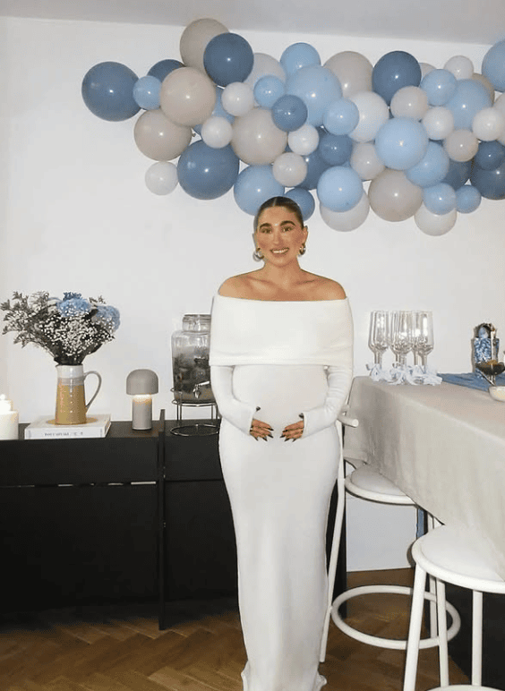 Elegant woman in white dress at baby shower with blue and beige balloons, celebrating a gender-neutr.