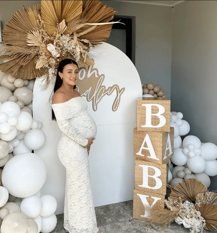 Pregnant woman smiling at baby shower with decorative balloons and floral arrangements.