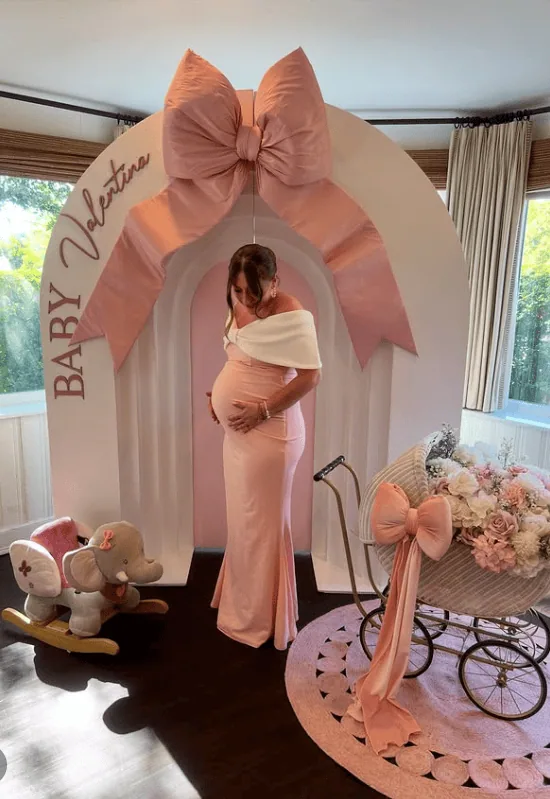 Elegant pregnant woman at baby shower with pink arch and floral decor.