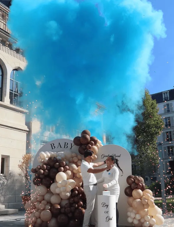 Couple celebrating gender reveal with colorful balloons and blue smoke explosion.