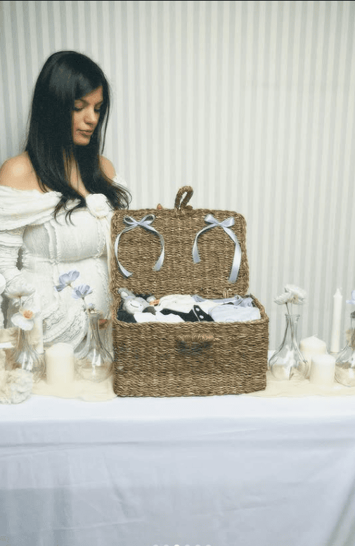 Woman with a baby shower gift basket filled with baby clothes and accessories on a table.