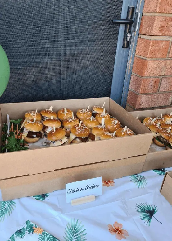 Assorted mini chicken sliders with buns and toppings in a cardboard box at a baby shower.