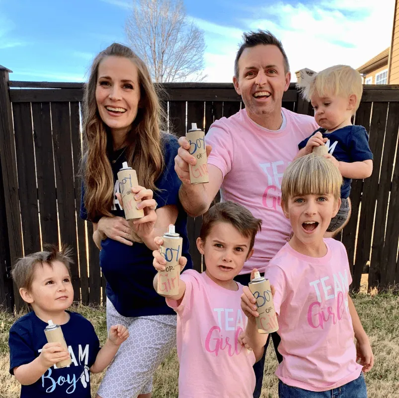 Family celebrating gender reveal with siblings holding decorated bottles outdoors.