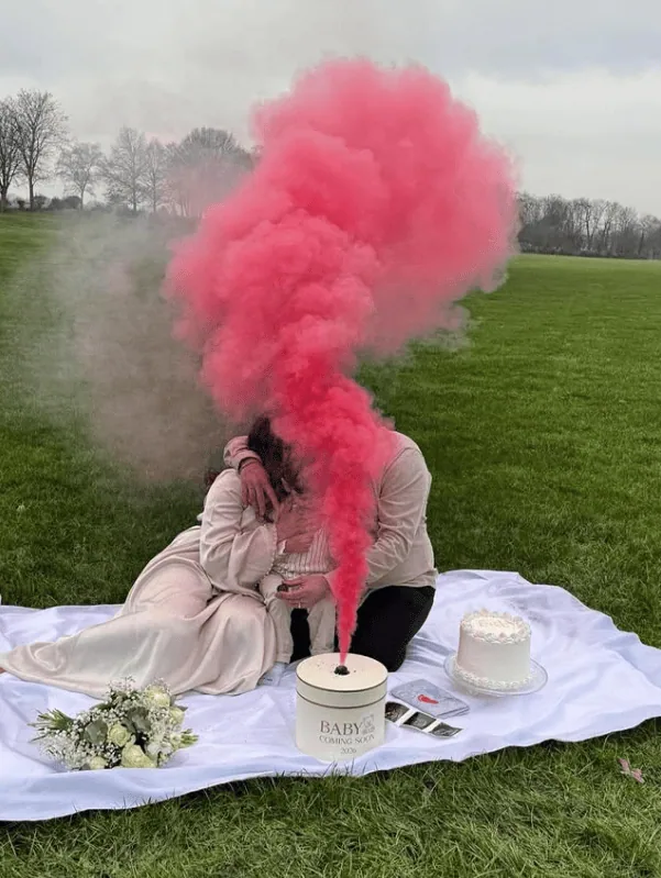Couple sharing a gender reveal moment with pink smoke in an outdoor park setting.