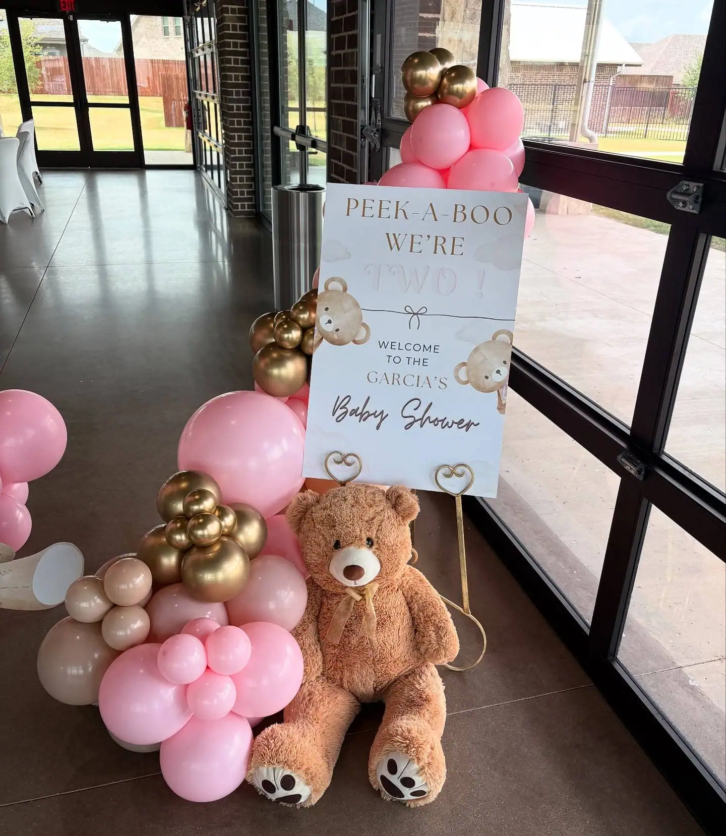 Baby shower welcome sign with pink and gold balloons, teddy bear, and decorative elements.