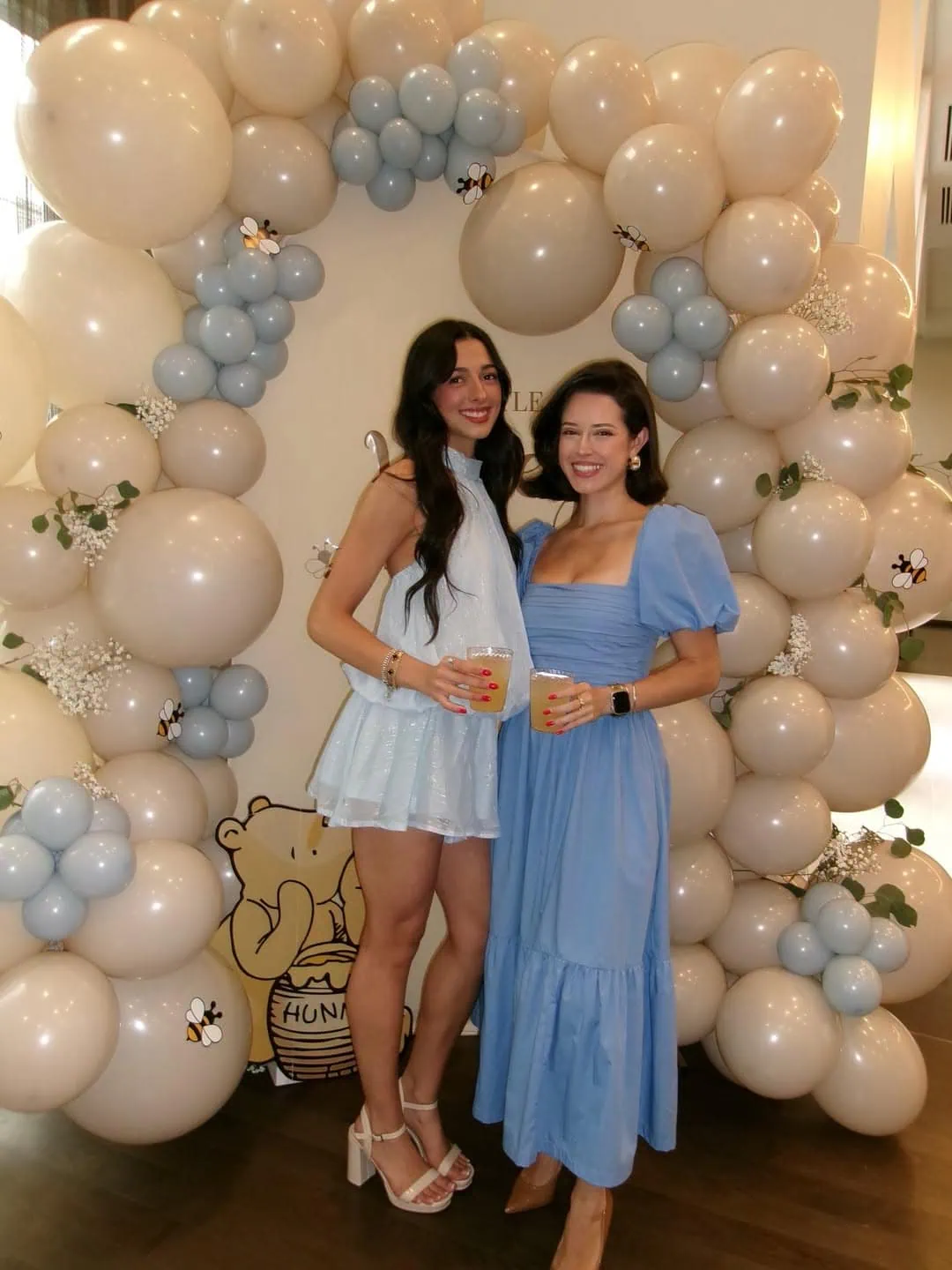 Two women in blue dresses at baby shower with balloon arch background.