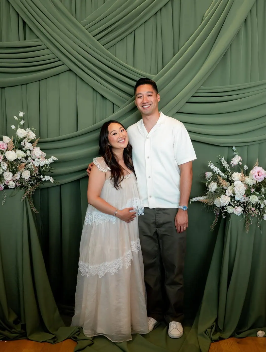 Smiling couple at a baby shower with elegant green backdrop and floral decor.
