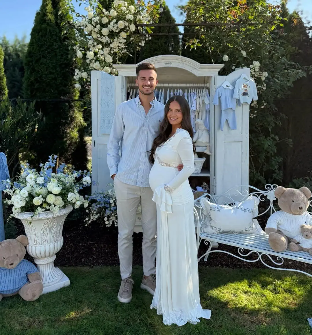 Pregnant woman in white dress with partner at outdoor baby shower with floral decor.