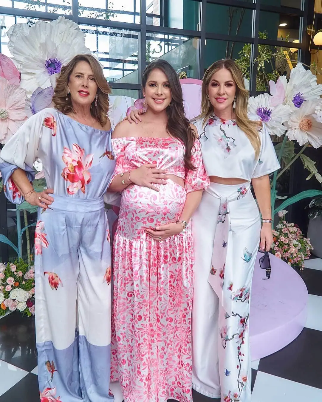 Three women in floral dresses celebrating at a baby shower with flowers and decorations.